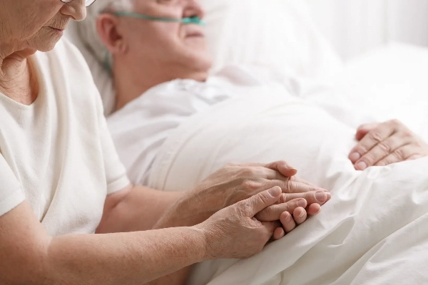 Elderly couple holding hands bedside, the wife staring at her husband's hands
