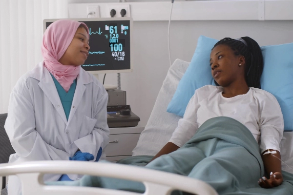 Smiling nurse wearing a hijab, sitting alongside a young patient laying in bed with a vital signs patient monitor in the background